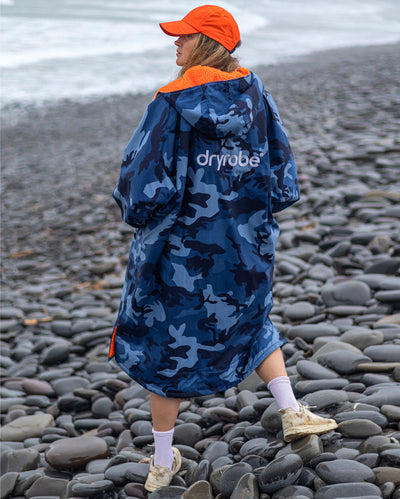 Woman stood on beach with back to camera, wearing Blue Camo Orange dryrobe advance long sleeve changing robe and orange cap 