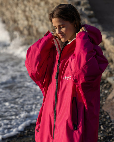 Woman stood in the sun on a beach, wearing dryrobe Advance long sleeve changing robe in Pink Light Grey