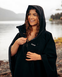 Close up of woman smiling wearing Black dryrobe® Waterproof Poncho, stood by a lake 