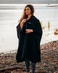 Woman stood by lake, wearing Black dryrobe® Waterproof Poncho with hood down