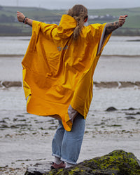Woman stood on a beach with back to the camera with arms in the air, wearing Yellow dryrobe® Waterproof Poncho 