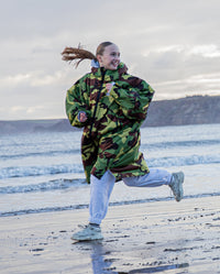 Girl smiling running along the shoreline, wearing Camo Grey dryrobe® Advance Kids Long Sleeve