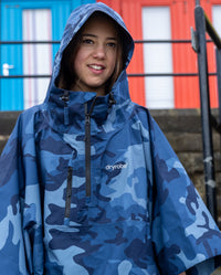 Girl stood in front of beach huts, wearing Blue Camo Kids dryrobe® Waterproof Poncho with hood up 