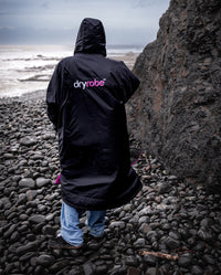 *MALE* stood on rocky beach with back to the camera, wearing Black Pink dryrobe® Advance Long Sleeve