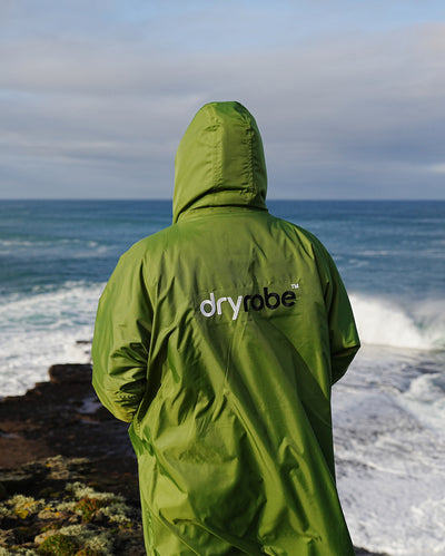 *MALE* Person stood facing the sea, wearing Forest Green dryrobe¬Æ Advance Long Sleeve with hood up