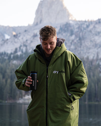 *MALE* stood in front of mountains, holding dryrobe® x Ocean Bottle Brew Flask and wearing Forest Green dryrobe® Advance Long Sleeve