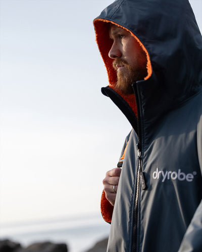 *MALE* Person wearing a dark Dryrobe with orange hood and 'dryrobe' branding, standing outdoors.
