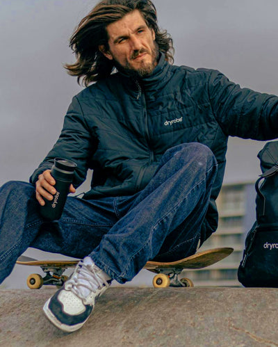 Man sat on skateboard, wearing dryrobe Mid-Layer Jacket in Black, holding dryrobe x Ocean Bottle Brew Flask
