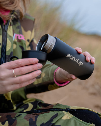 Person sat in sand dunes, pouring contents of dryrobe Ocean Bottle into lid
