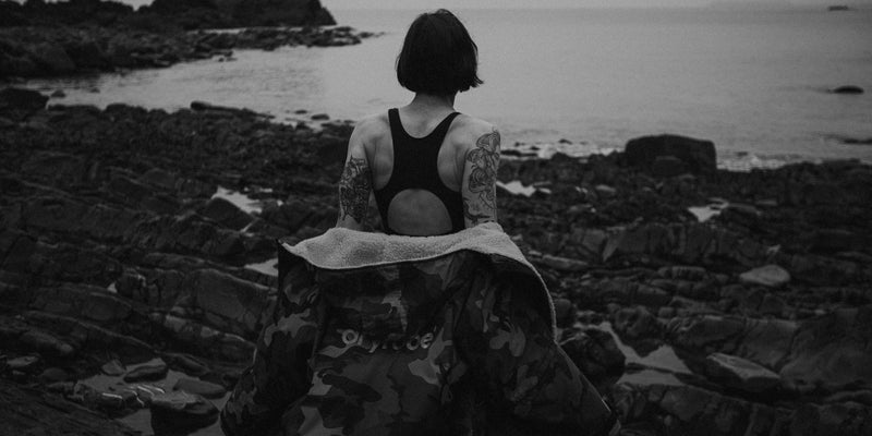 Person sitting on rocks by the ocean with tattoos visible