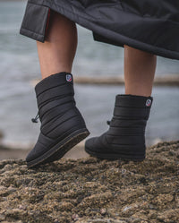 1|Close up of  dryrobe® Eco Thermal Boots being worn on a beach, with one heel raised up
