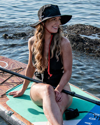 1|Woman sat on a paddle board in the sea, wearing Black dryrobe® Quick Dry brimmed hat